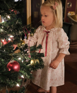 Girls' Nightie in Cordelia Toile, child in the pink toile print with red ribbon tie looking at baubles on a Christmas tree