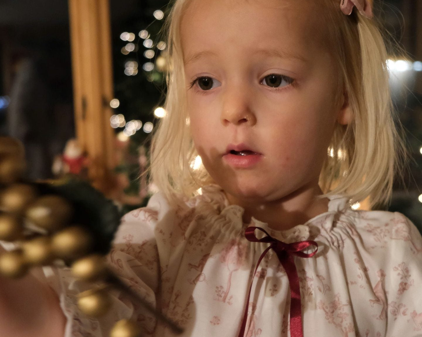 Girls' Nightie in Cordelia Toile, close-up of the gathered neckline and red ribbon bow on a child holding gold baubles