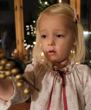 Girls' Nightie in Cordelia Toile, close-up of the gathered neckline and red ribbon bow on a child holding gold baubles