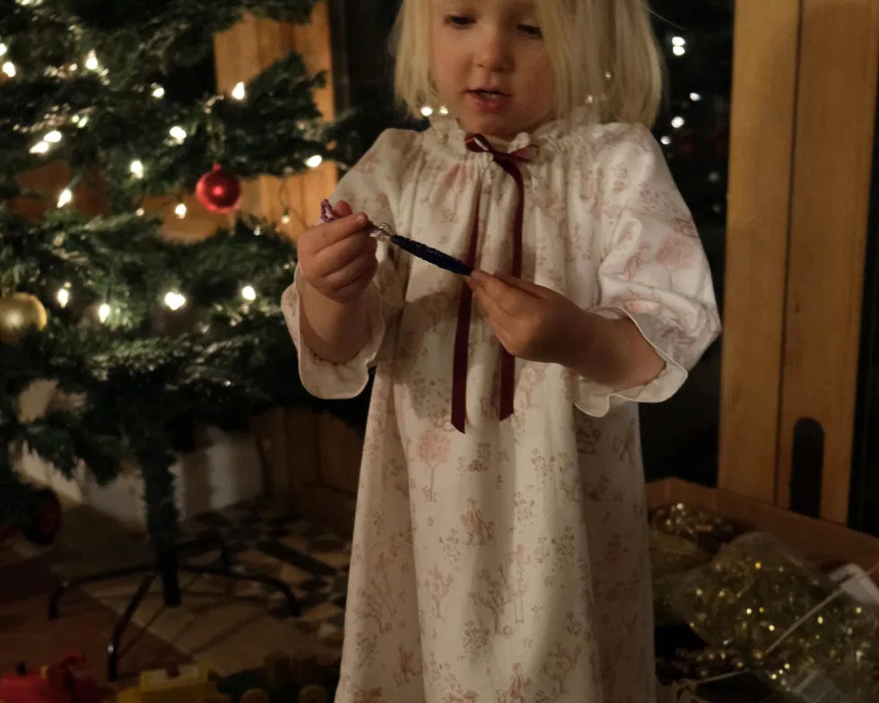Girls' Nightie in Cordelia Toile, full-length view on a child holding a small ornament beside a lit Christmas tree
