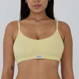 A woman wearing a pale yellow ribbed bralette with thin straps and a small white brand label that reads 'RENUE' at the center band, shown against a plain light gray background.