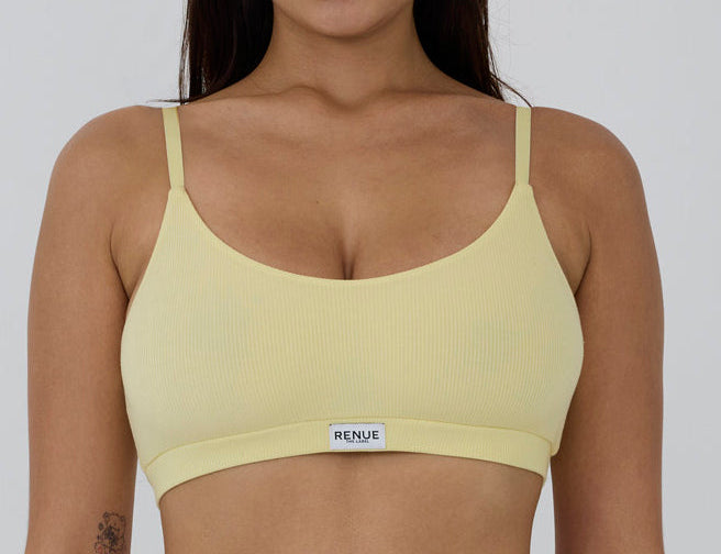 A woman wearing a pale yellow ribbed bralette with thin straps and a small white brand label that reads 'RENUE' at the center band, shown against a plain light gray background.