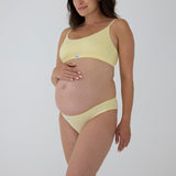 A pregnant model wearing a light yellow maternity bra and matching underwear set against a plain light gray background.