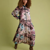 A woman modeling a long-sleeved midi dress with a floral print in dusty pink, teal, and brown tones, paired with brown pointed-toe heels, against a solid olive green background.