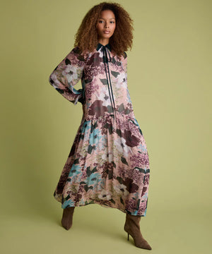 A woman modeling a long-sleeved midi dress with a floral print in dusty pink, teal, and brown tones, paired with brown pointed-toe heels, against a solid olive green background.