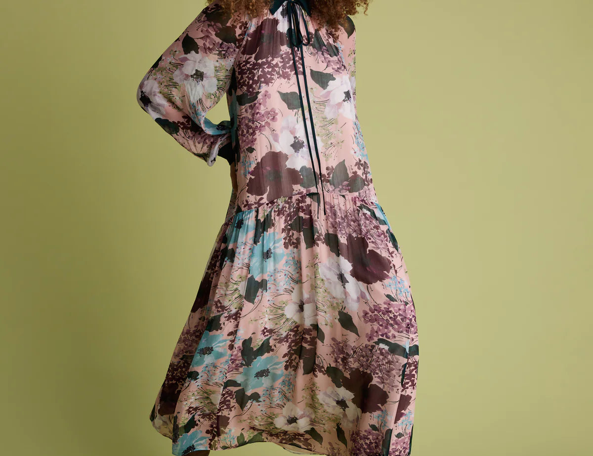 A woman modeling a long-sleeved midi dress with a floral print in dusty pink, teal, and brown tones, paired with brown pointed-toe heels, against a solid olive green background.