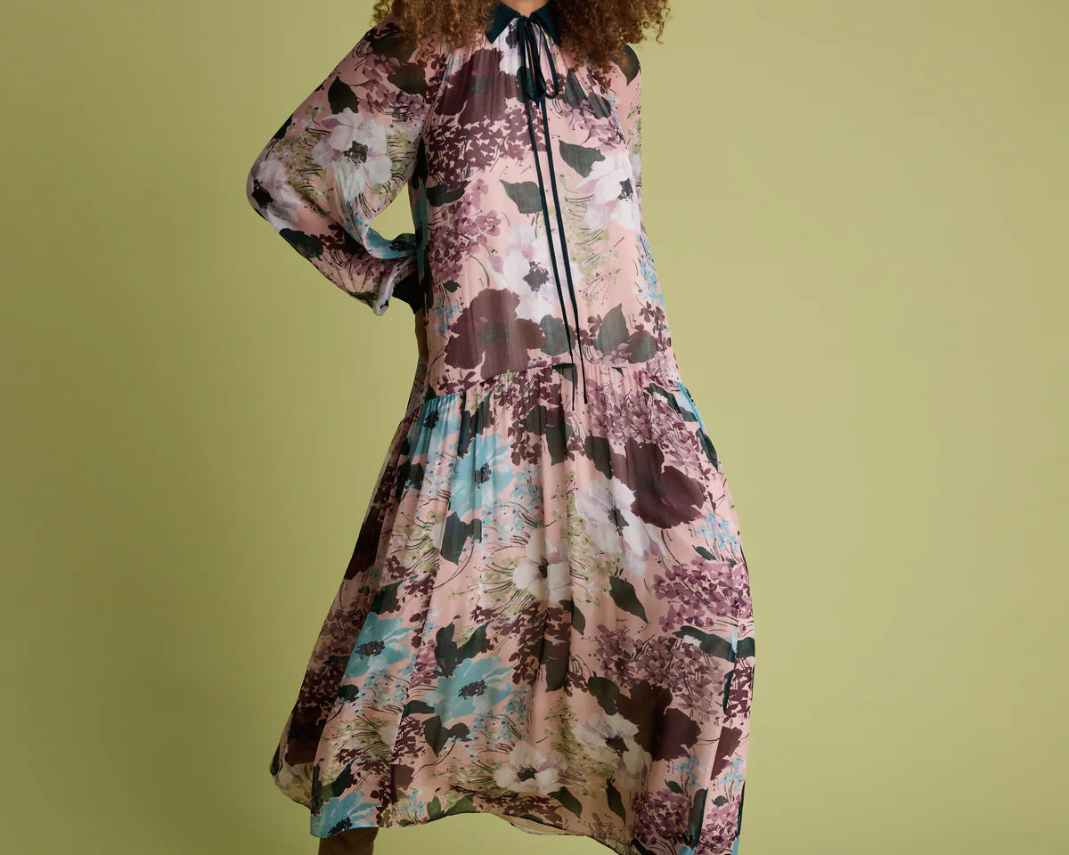 A woman modeling a long-sleeved midi dress with a floral print in dusty pink, teal, and brown tones, paired with brown pointed-toe heels, against a solid olive green background.