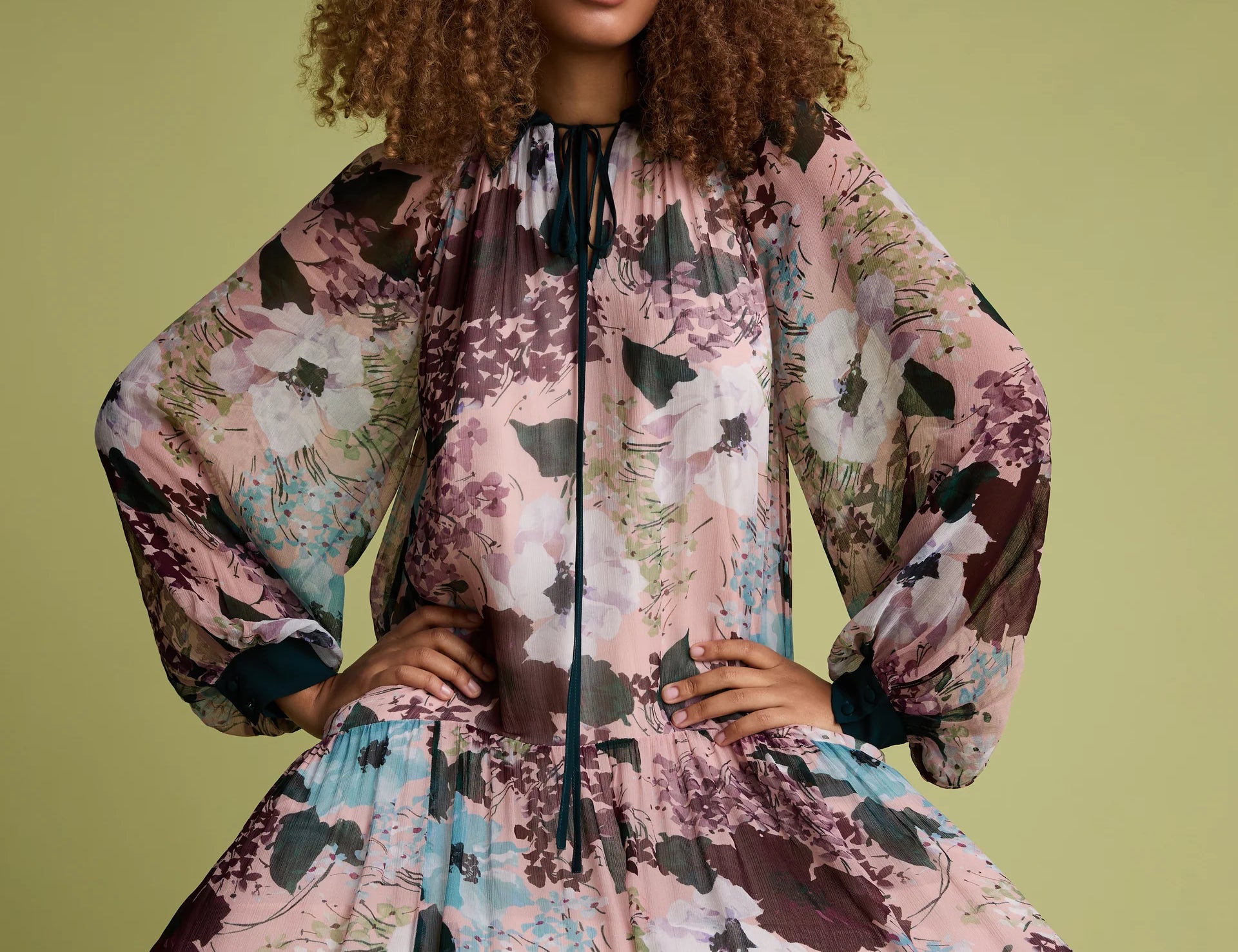 A model with curly brown hair wearing a voluminous, long-sleeved floral dress with a high neck and black tie detail, posed against a solid olive green background.