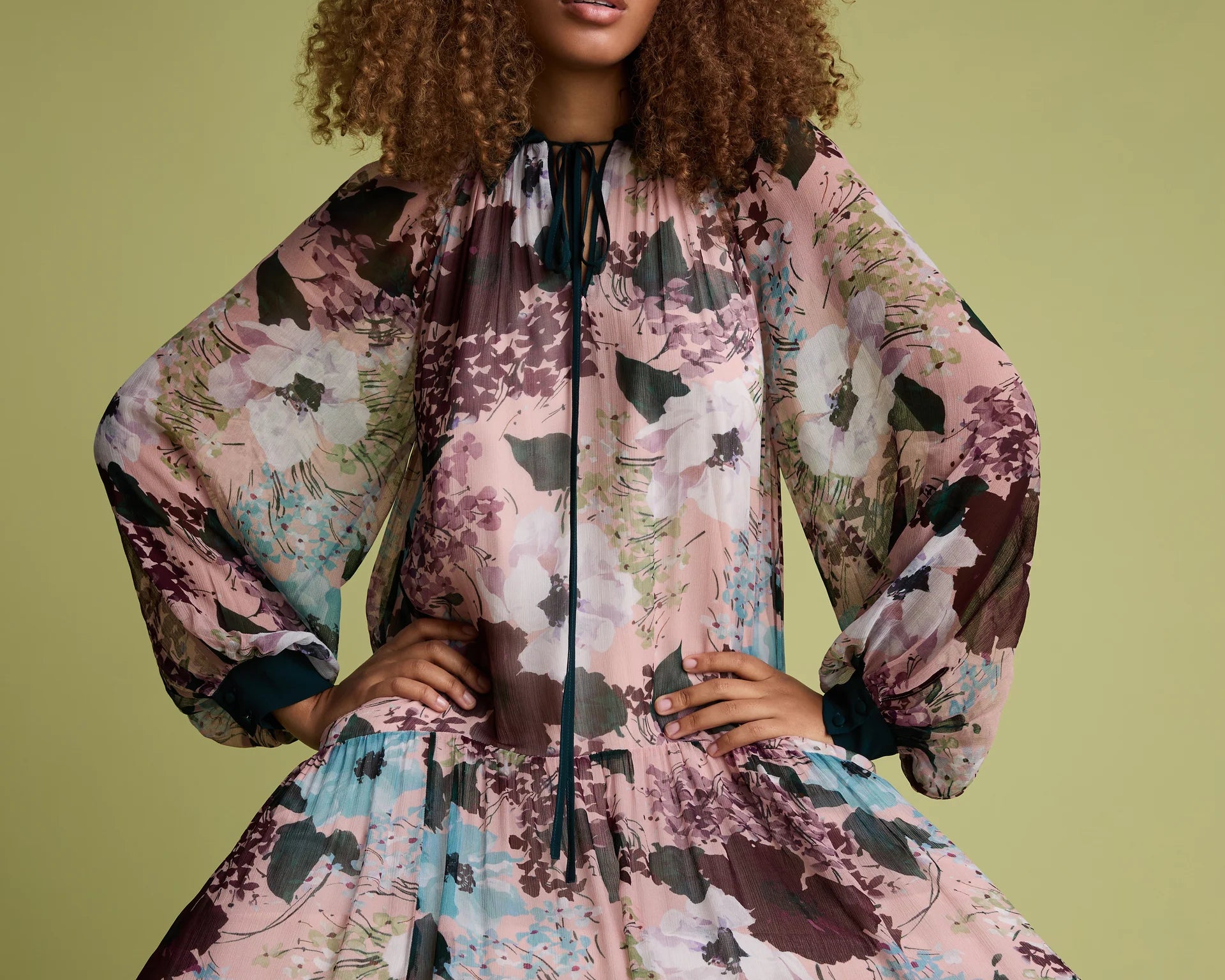 A model with curly brown hair wearing a voluminous, long-sleeved floral dress with a high neck and black tie detail, posed against a solid olive green background.