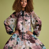 A model with curly brown hair wearing a voluminous, long-sleeved floral dress with a high neck and black tie detail, posed against a solid olive green background.