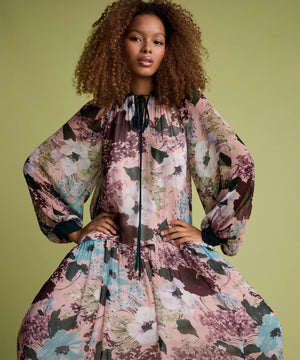 A model with curly brown hair wearing a voluminous, long-sleeved floral dress with a high neck and black tie detail, posed against a solid olive green background.