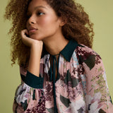 A woman with curly brown hair wearing a pink floral long-sleeve blouse with dark teal trim, resting her chin on her hand against a solid olive green background.