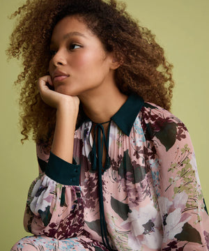 A woman with curly brown hair wearing a pink floral long-sleeve blouse with dark teal trim, resting her chin on her hand against a solid olive green background.