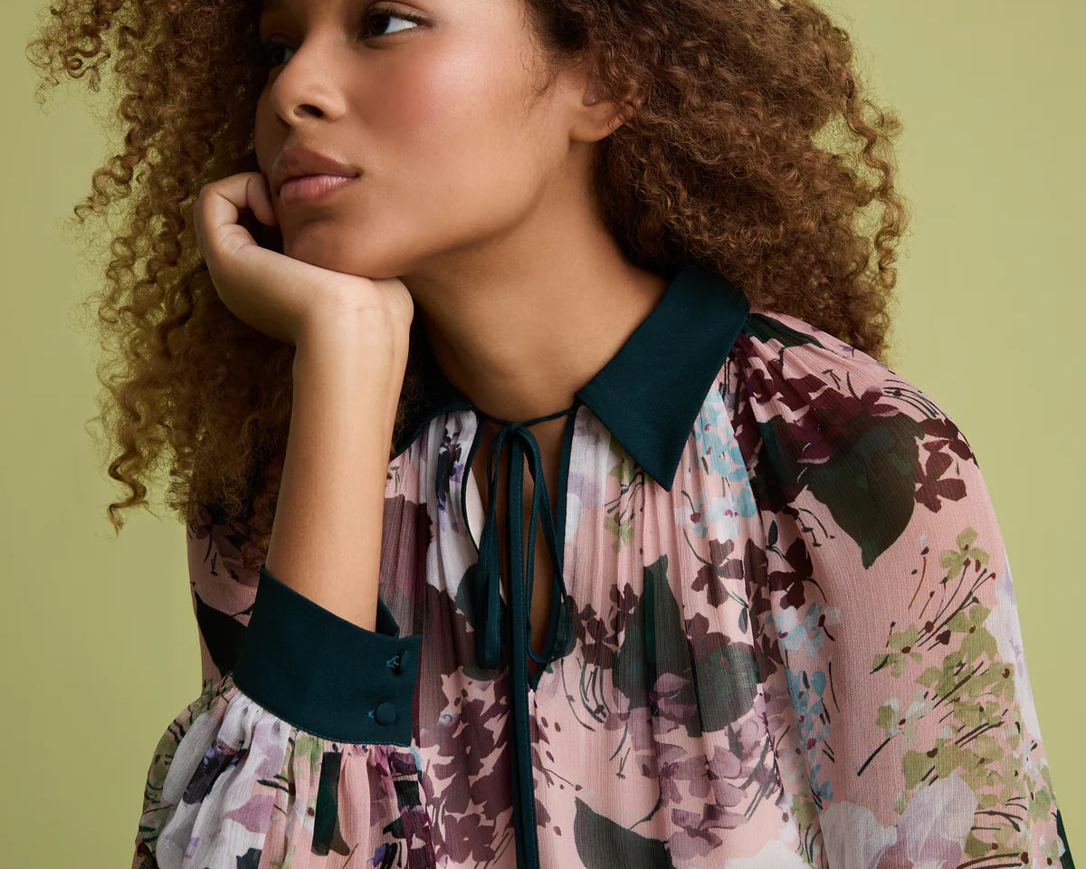 A woman with curly brown hair wearing a pink floral long-sleeve blouse with dark teal trim, resting her chin on her hand against a solid olive green background.