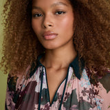 A close-up portrait of a woman with voluminous curly brown hair wearing a sheer floral blouse with a dark teal collar and trim. The blouse features a pink, purple, and green floral pattern.