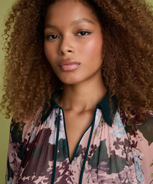 A close-up portrait of a woman with voluminous curly brown hair wearing a sheer floral blouse with a dark teal collar and trim. The blouse features a pink, purple, and green floral pattern.