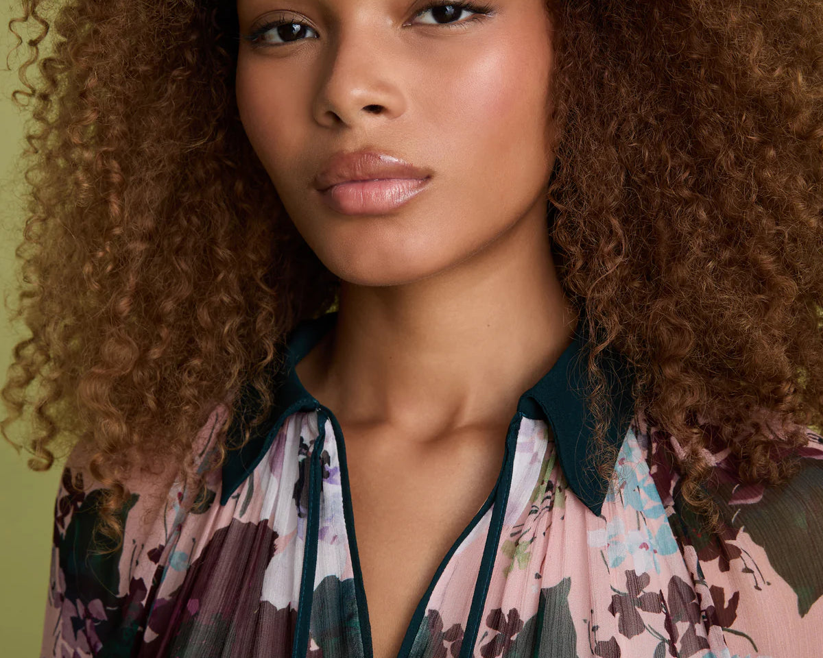 A close-up portrait of a woman with voluminous curly brown hair wearing a sheer floral blouse with a dark teal collar and trim. The blouse features a pink, purple, and green floral pattern.