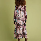 A woman viewed from behind wearing a long-sleeved maxi dress with a pink, brown, and teal abstract floral print, paired with brown ankle boots, set against a solid olive green background.