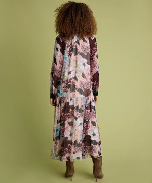 A woman viewed from behind wearing a long-sleeved maxi dress with a pink, brown, and teal abstract floral print, paired with brown ankle boots, set against a solid olive green background.