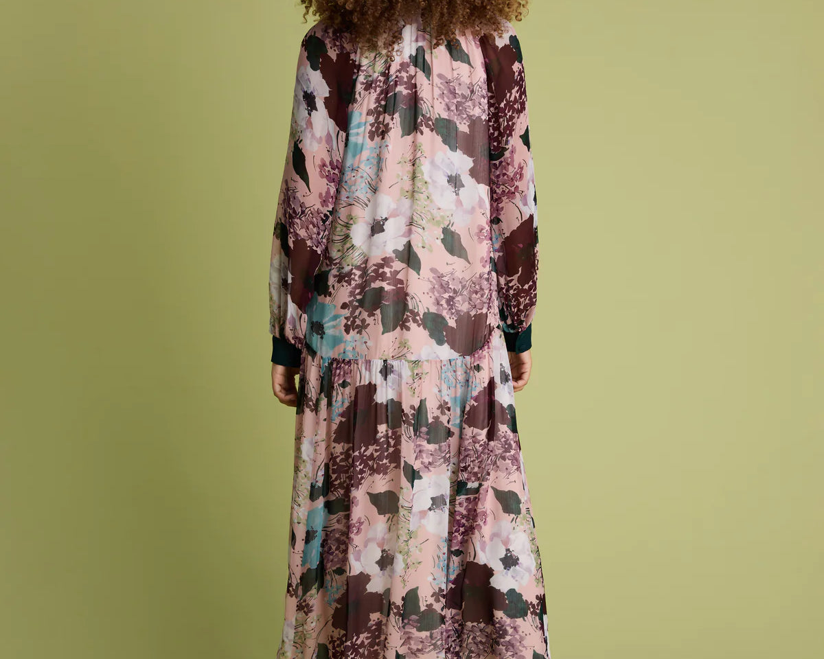 A woman viewed from behind wearing a long-sleeved maxi dress with a pink, brown, and teal abstract floral print, paired with brown ankle boots, set against a solid olive green background.