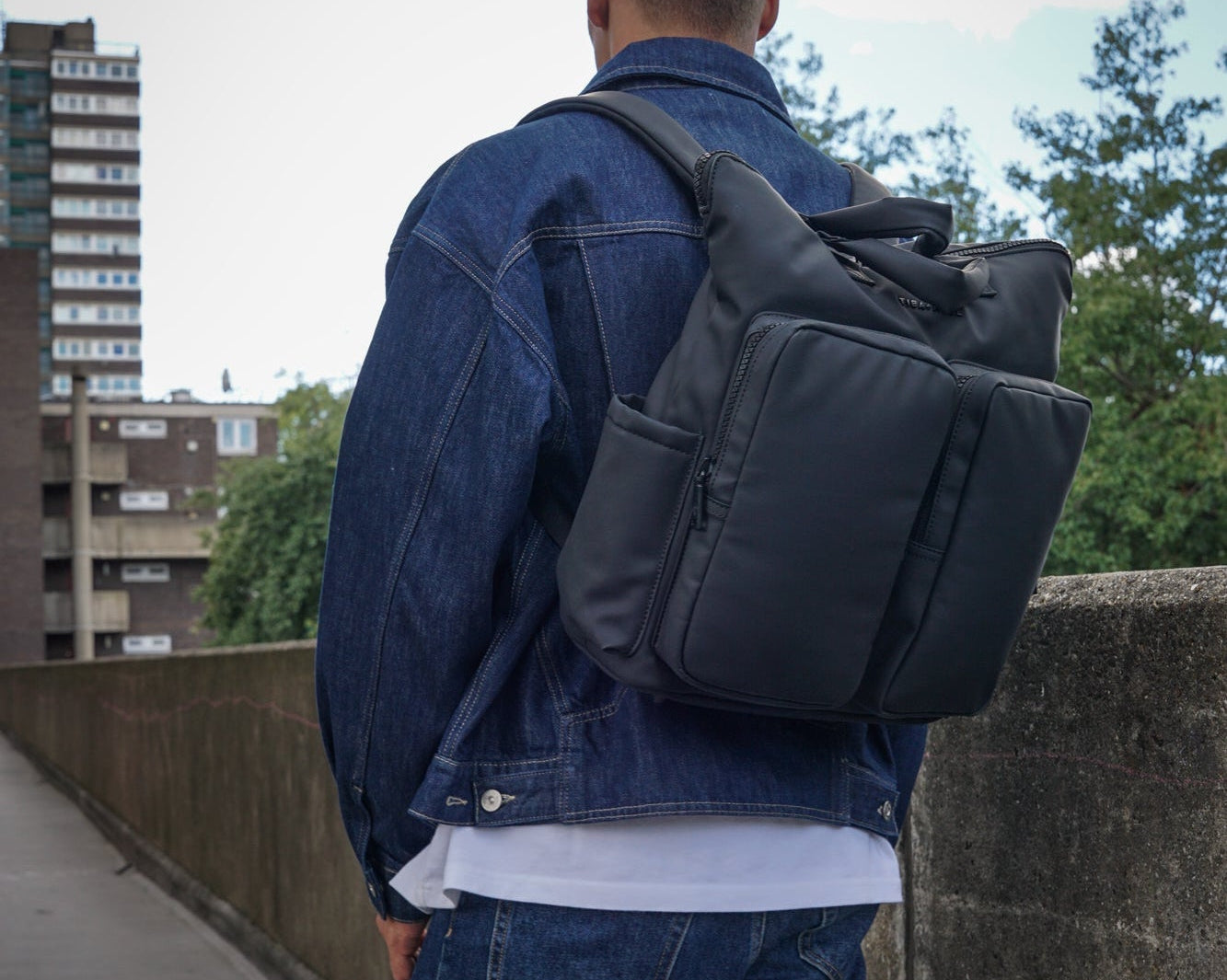 Person wearing a black backpack and denim jacket on a bridge with buildings and trees in the background