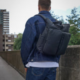 Person wearing a black backpack and denim jacket on a bridge with buildings and trees in the background