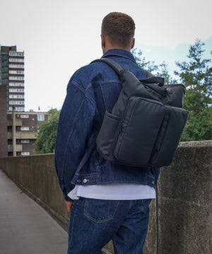Person wearing a black backpack and denim jacket on a bridge with buildings and trees in the background