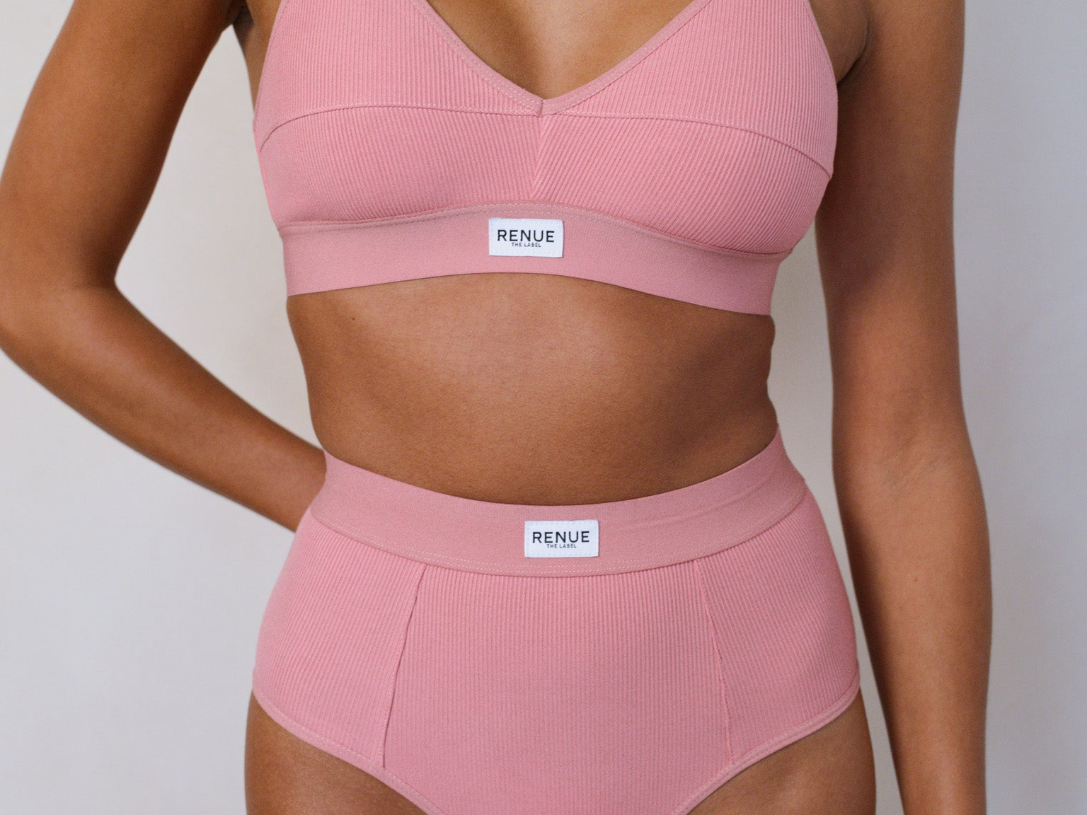 A model wearing a two-piece ribbed pink lingerie set consisting of a V-neck bralette and matching high-waisted briefs, both featuring a white rectangular 'RENUE' brand label on the waistband.