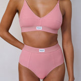 A model wearing a two-piece ribbed pink lingerie set consisting of a V-neck bralette and matching high-waisted briefs, both featuring a white rectangular 'RENUE' brand label on the waistband.