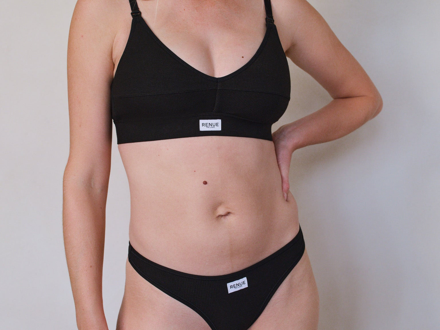 A woman modeling a black two-piece bikini set against a plain light gray wall. The set includes a black V-neck bikini top with thin straps and a matching black bikini bottom with a small white brand label.