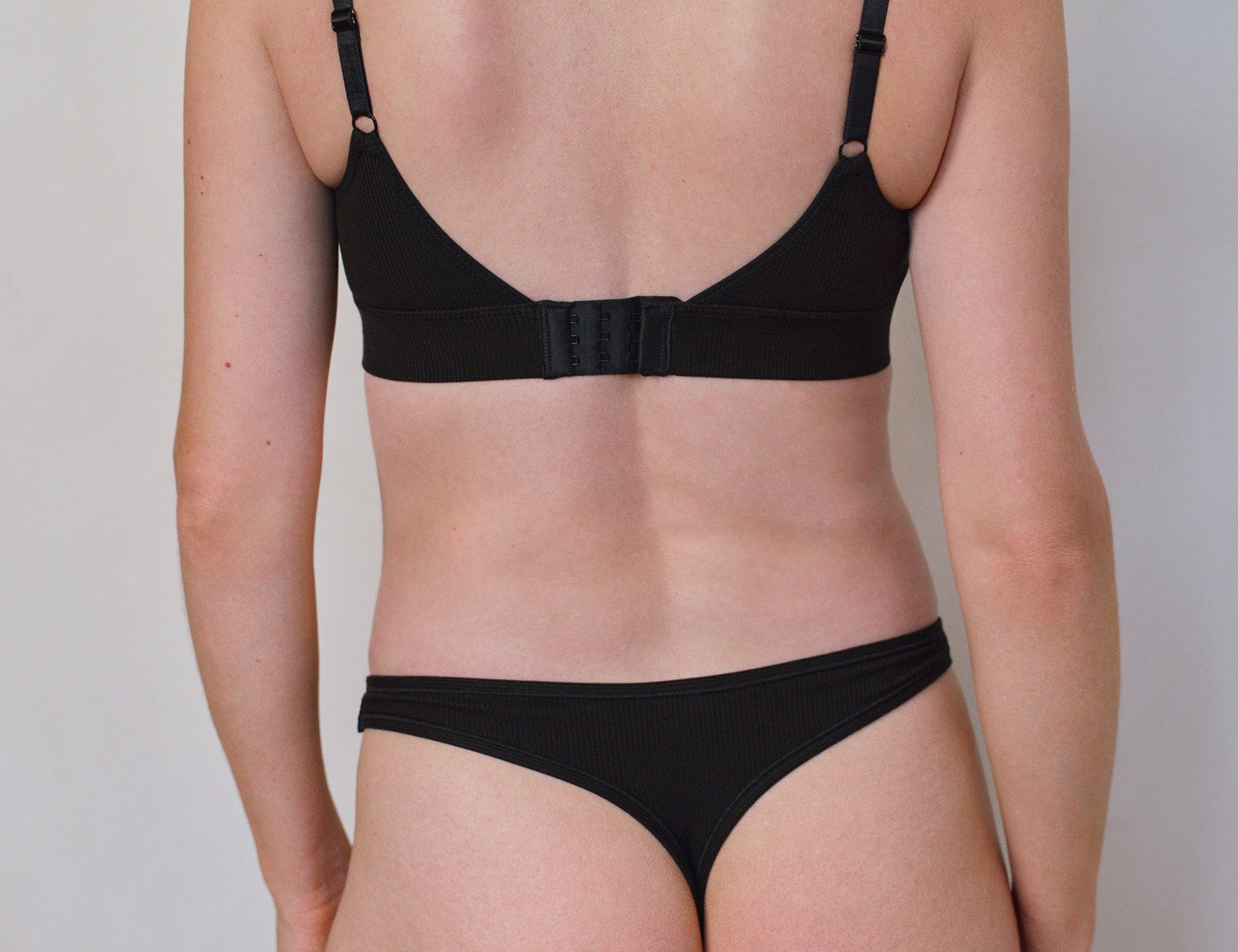 Rear view of a model wearing a black two-piece lingerie set consisting of a bralette with adjustable shoulder straps and a matching thong, against a plain light gray background.