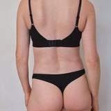 Rear view of a model wearing a black two-piece lingerie set consisting of a bralette with adjustable shoulder straps and a matching thong, against a plain light gray background.