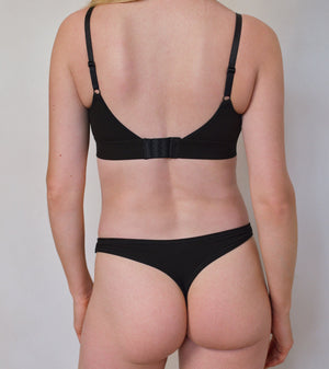 Rear view of a model wearing a black two-piece lingerie set consisting of a bralette with adjustable shoulder straps and a matching thong, against a plain light gray background.