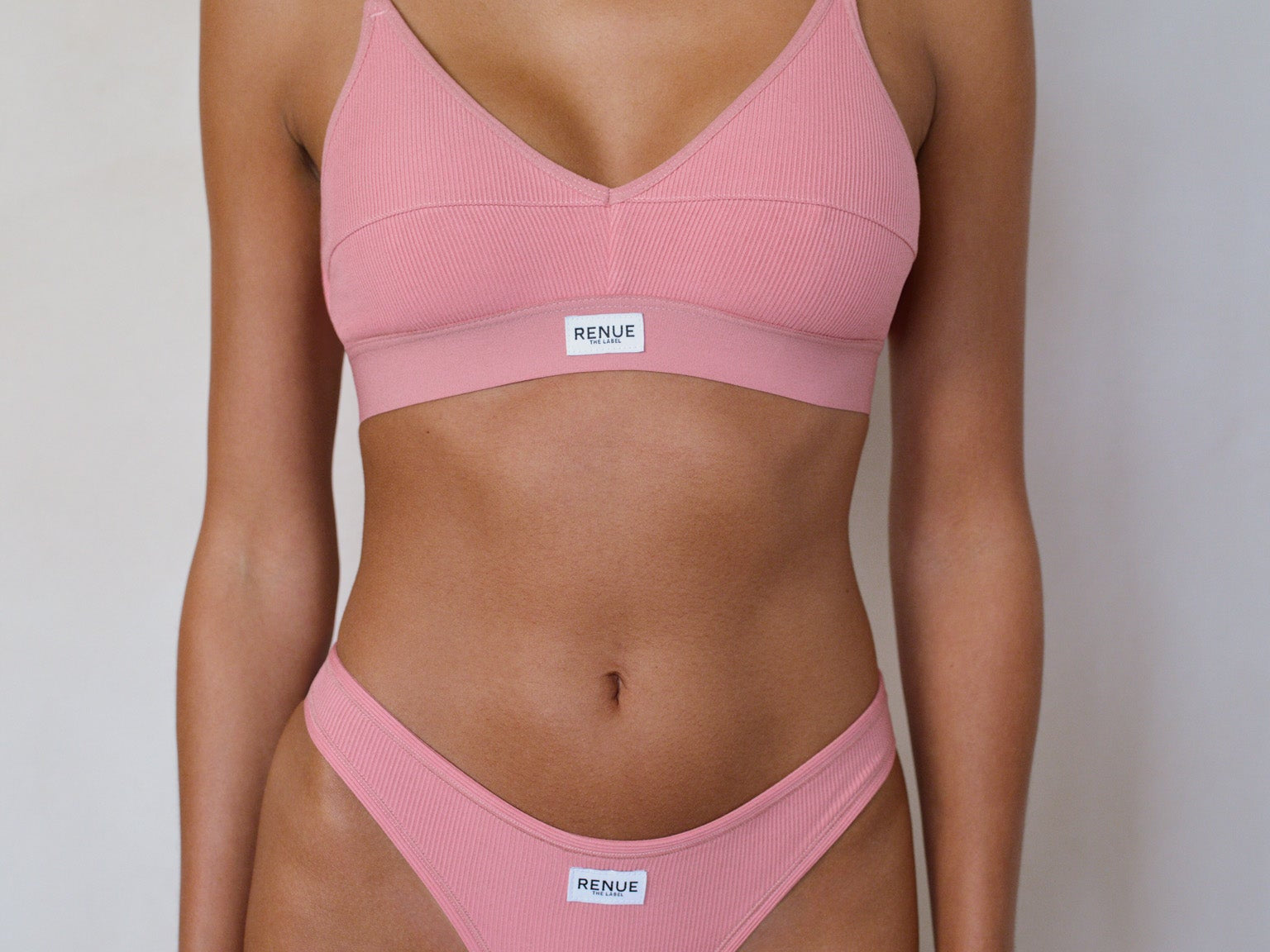 A model wearing a dusty pink two-piece bikini set with thin straps and a small white brand label on the front of each piece, set against a plain light gray background.