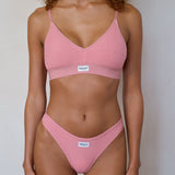 A model wearing a dusty pink two-piece bikini set with thin straps and a small white brand label on the front of each piece, set against a plain light gray background.