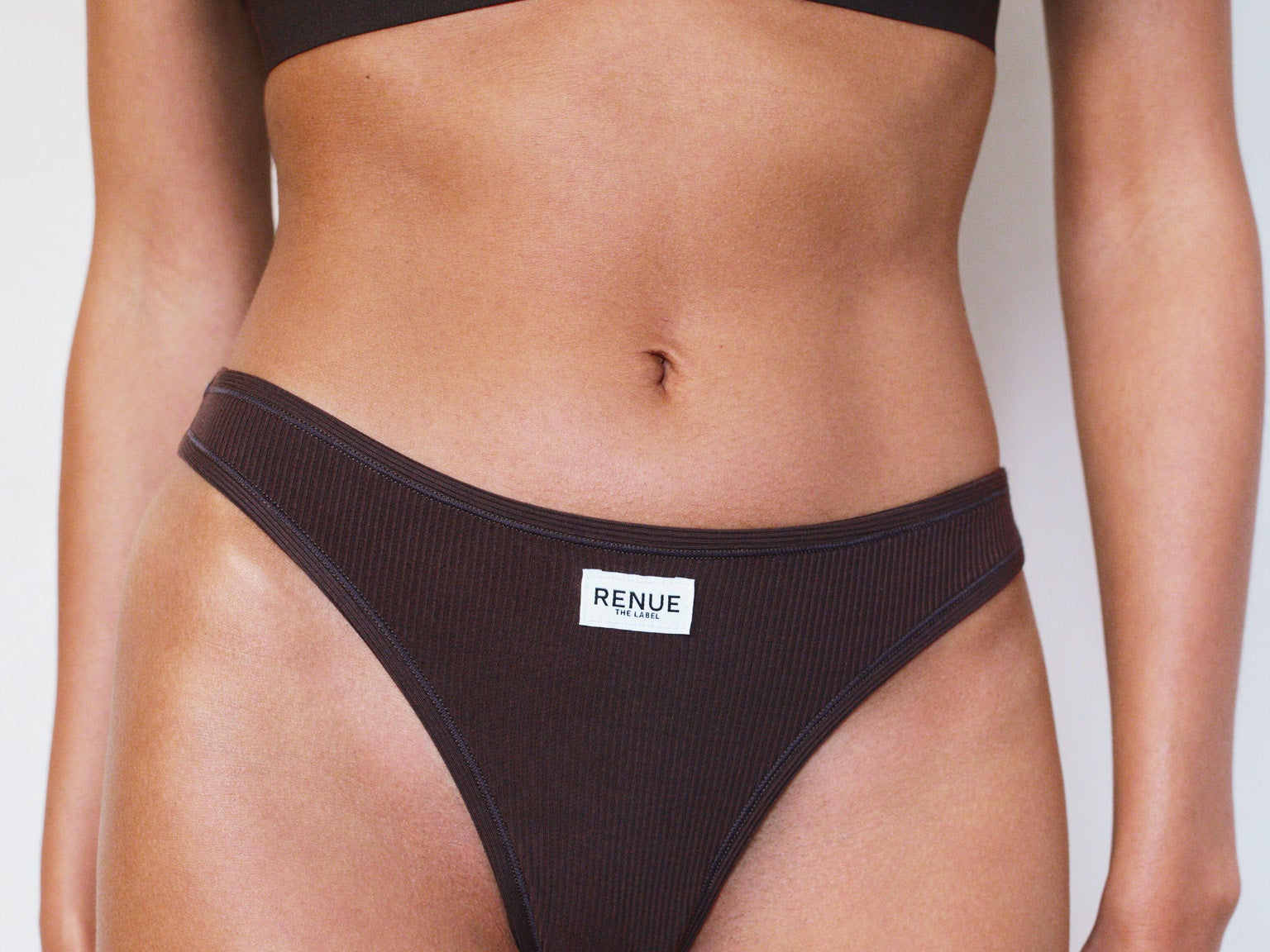 A model wearing a ribbed two-piece bikini set in dark brown, featuring a matching sports bra top and high-cut bikini bottoms with a small white brand label reading 'RENUE'.