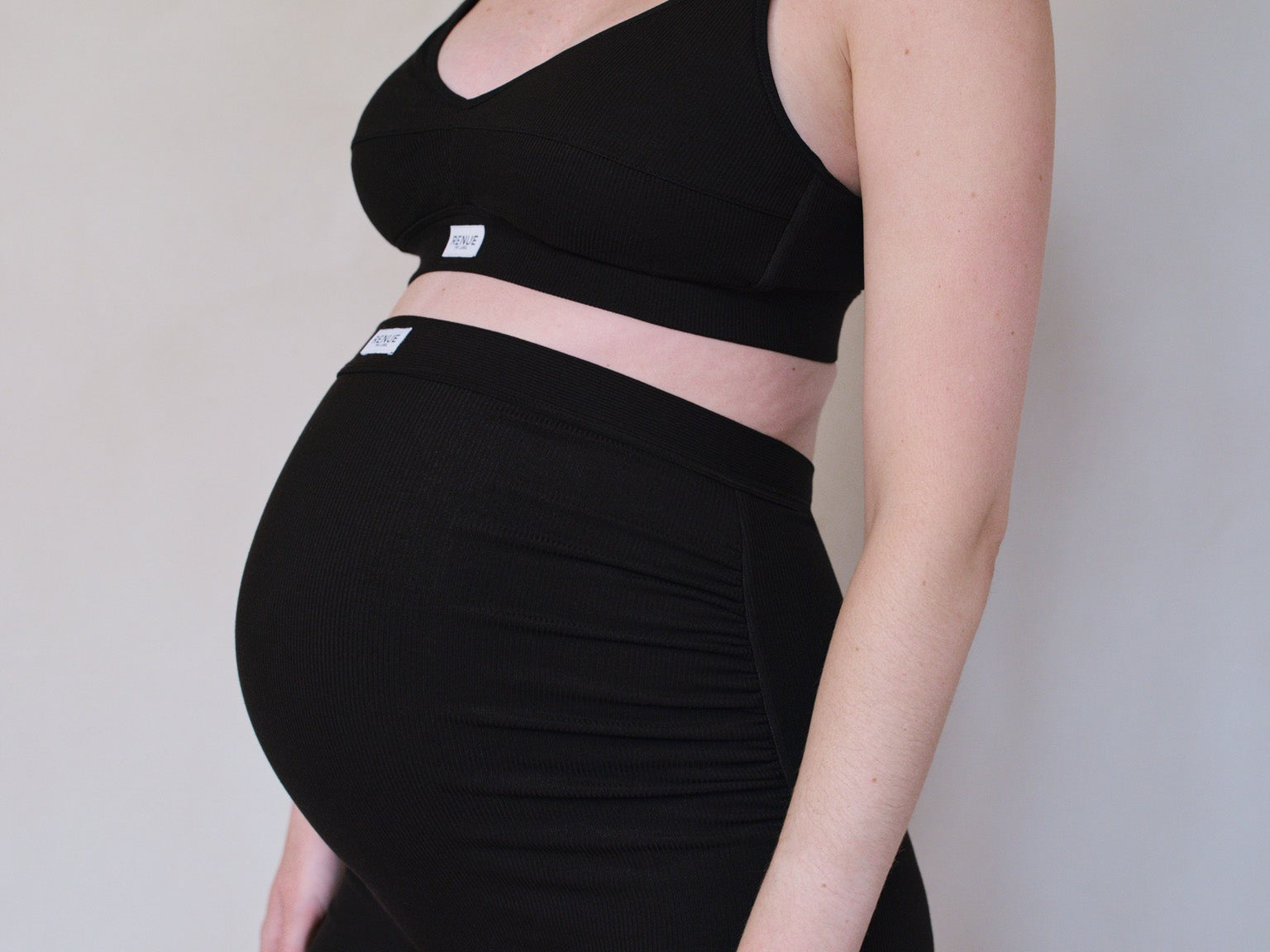 A pregnant model wearing a matching black two-piece activewear set consisting of a cropped tank top and high-waisted ruched shorts, shown from the side against a plain light beige wall.