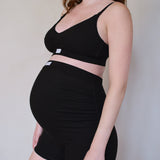 A pregnant model wearing a matching black two-piece activewear set consisting of a cropped tank top and high-waisted ruched shorts, shown from the side against a plain light beige wall.
