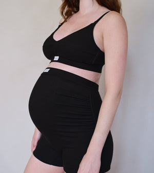 A pregnant model wearing a matching black two-piece activewear set consisting of a cropped tank top and high-waisted ruched shorts, shown from the side against a plain light beige wall.