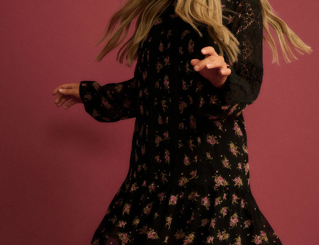 A woman wearing a black long-sleeve mini dress with a small pink and green floral print, photographed from behind against a solid dusty rose background.
