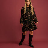 A woman modeling a black long-sleeve mini dress with a small floral pattern, paired with black knee-high boots, set against a solid dusty rose studio backdrop.
