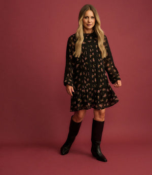 A woman modeling a black long-sleeve mini dress with a small floral pattern, paired with black knee-high boots, set against a solid dusty rose studio backdrop.