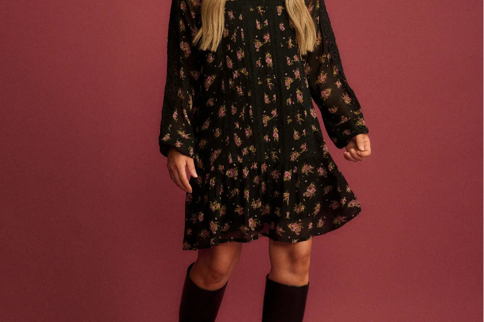 A woman modeling a black long-sleeve mini dress with a small floral pattern, paired with black knee-high boots, set against a solid dusty rose studio backdrop.