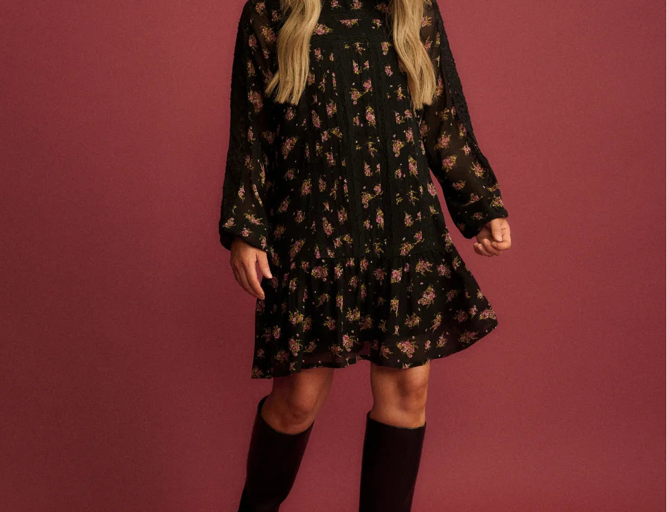 A woman modeling a black long-sleeve mini dress with a small floral pattern, paired with black knee-high boots, set against a solid dusty rose studio backdrop.