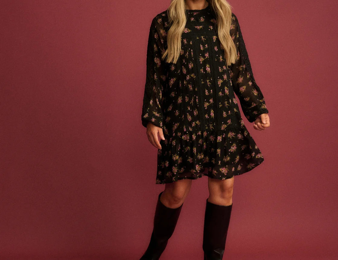 A woman modeling a black long-sleeve mini dress with a small floral pattern, paired with black knee-high boots, set against a solid dusty rose studio backdrop.