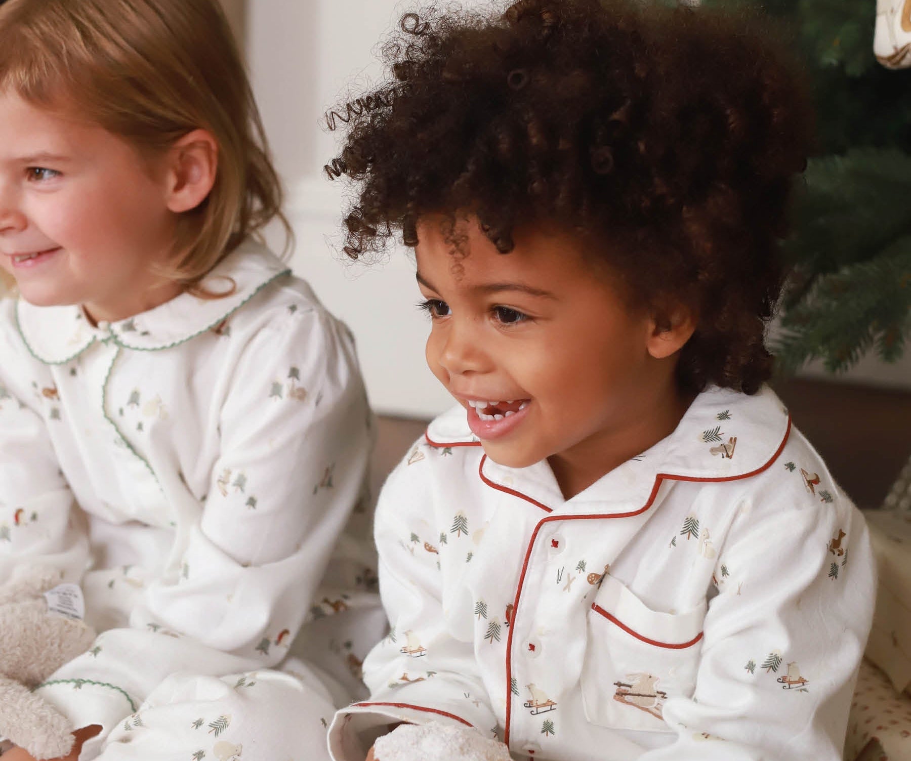 Two children in white pajamas with red trim sitting next to a Christmas tree.
