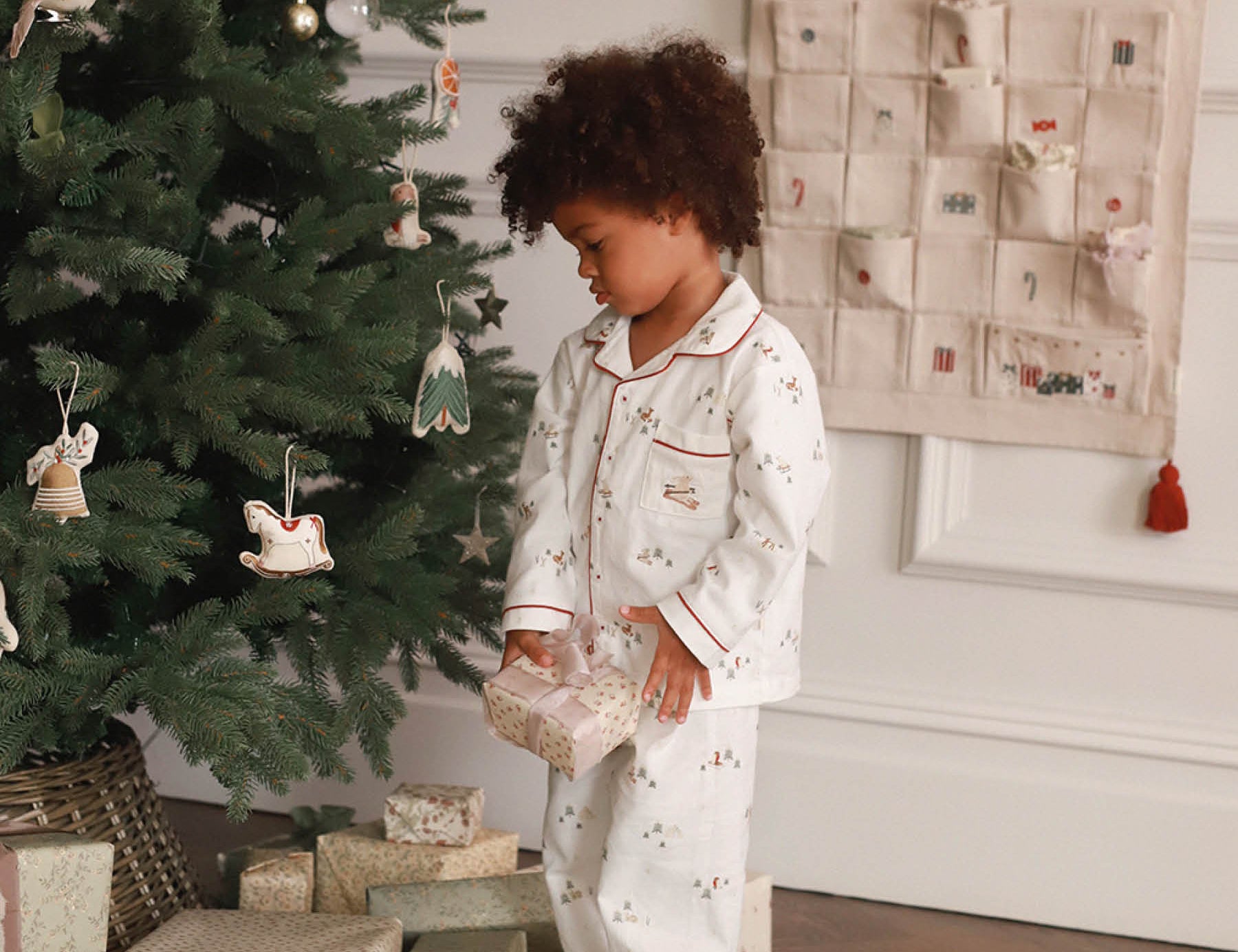 Child in pajamas standing next to a decorated Christmas tree with presents underneath.