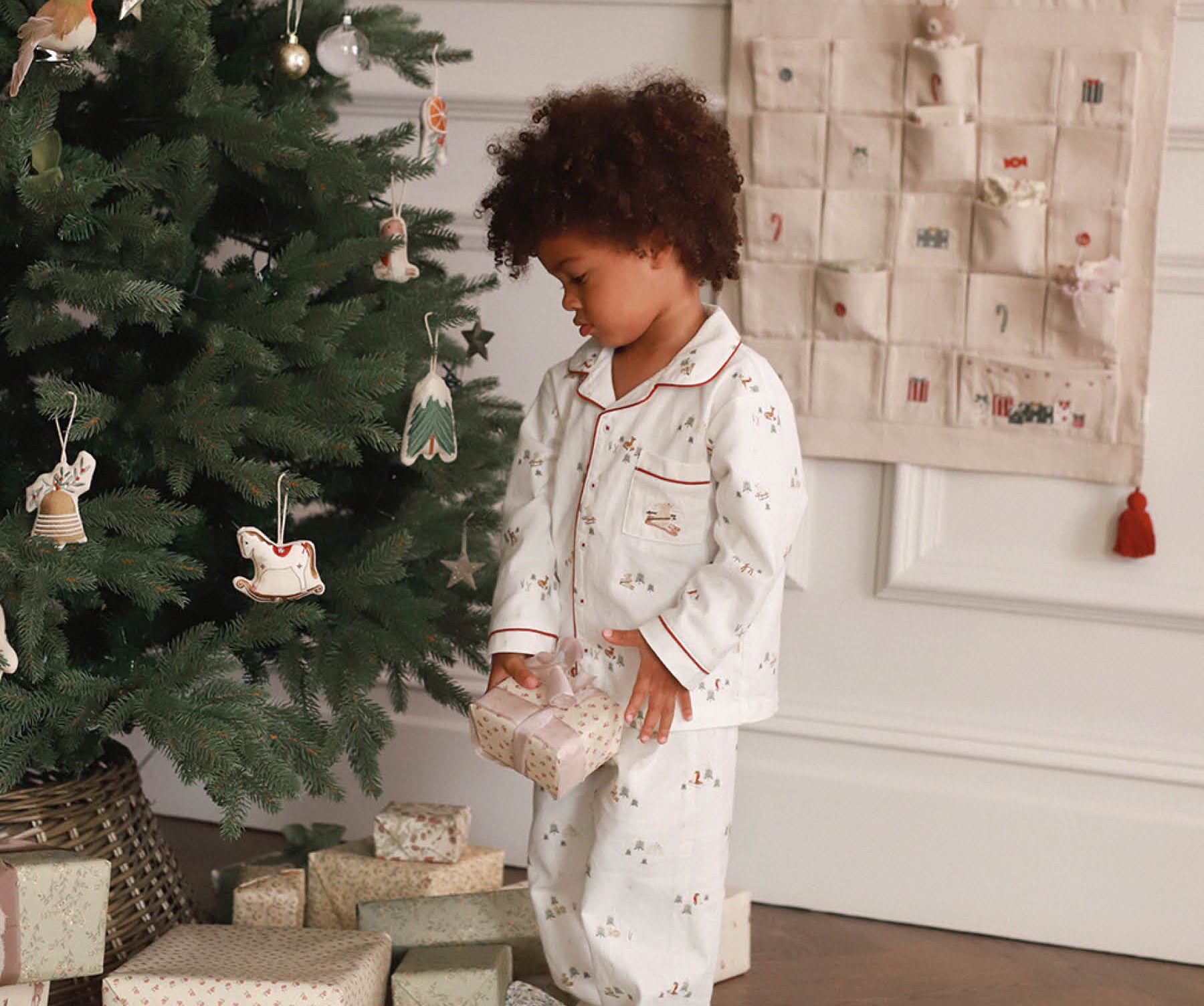 Child in pajamas standing next to a decorated Christmas tree with presents underneath.