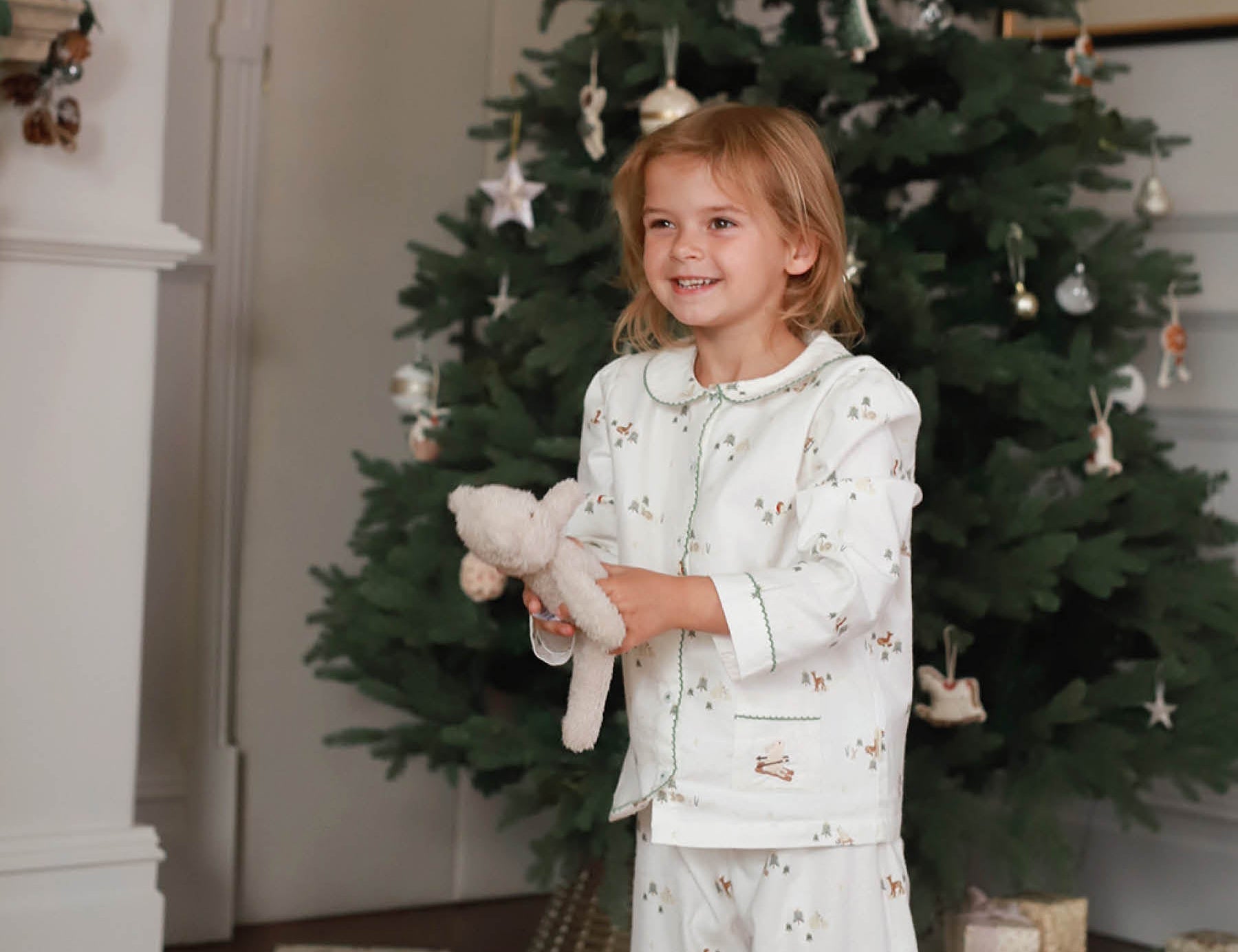 Child in pajamas holding a teddy bear in front of a decorated Christmas tree.