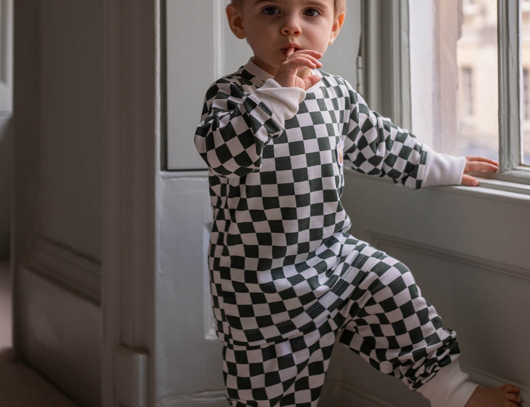 Child wearing a black and white checkered outfit standing by a window.